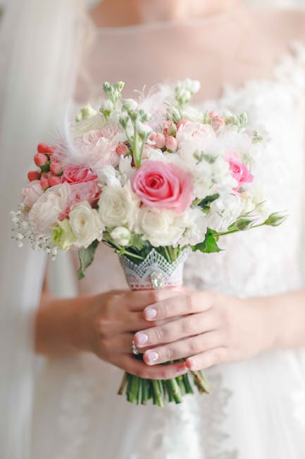 A bouquet of fresh flowers placed on a crumpled white bedsheet, featuring pink gerbera daisies and white daisy-like flowers with dark centers, arranged with green foliage. The bouquet is wrapped in br
