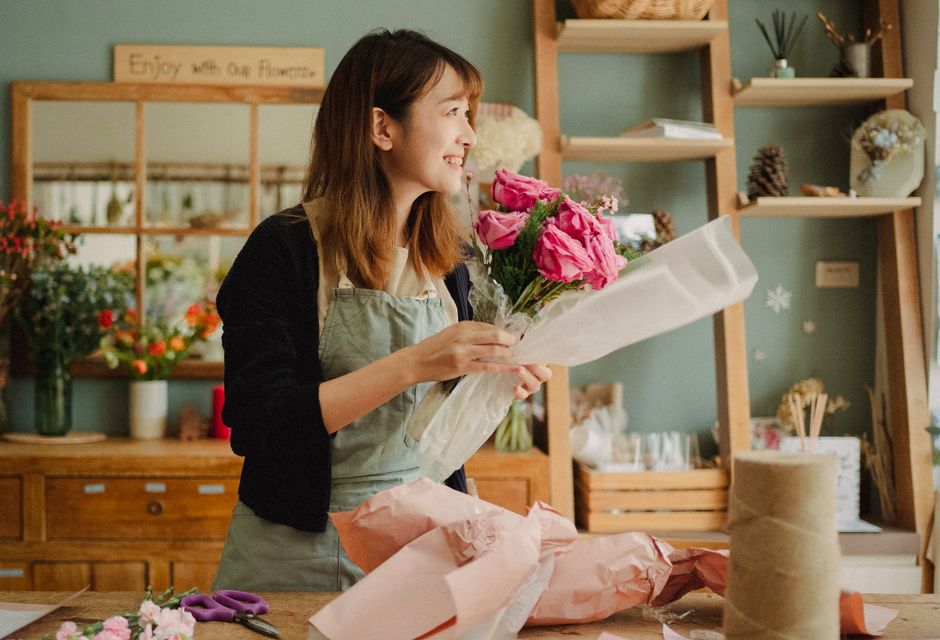 A young woman with long brown hair and light skin, dressed in a beige cardigan, white top, and black polka-dot skirt, is arranging a bouquet of white and pink flowers in a florist shop. The shop has a