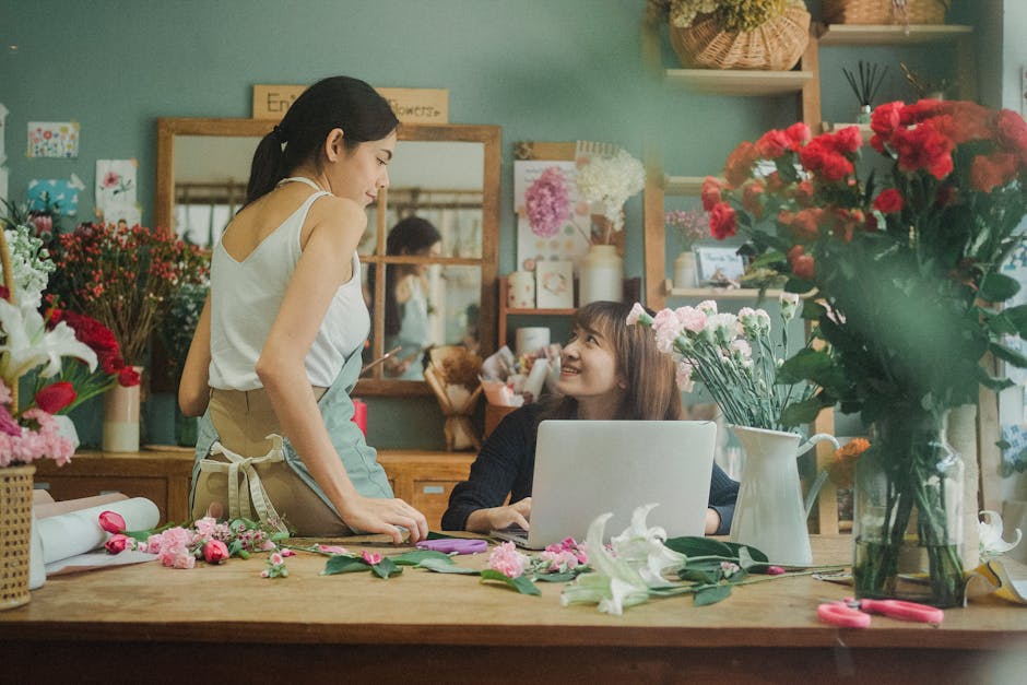 A young woman with long dark hair, smiling as she converses on a smartphone, stands behind a table with a floral arrangement. The bouquet includes white tulips with green stems, accented with red and 