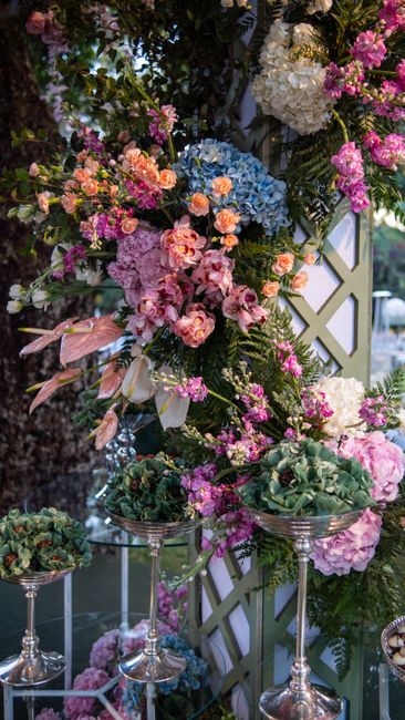 An arrangement of fresh flowers featuring pastel pink roses, blue hydrangeas, and white peonies, complemented by sprigs of purple and pink blossoms. The flowers are arranged in a cascading style, disp