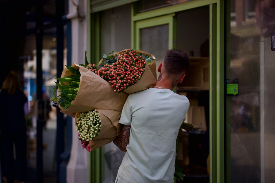 A young woman with long dark hair, wearing a white t-shirt and black apron, is standing in a floral shop with a welcoming smile. She is holding a clear glass vase filled with fresh yellow tulips and w