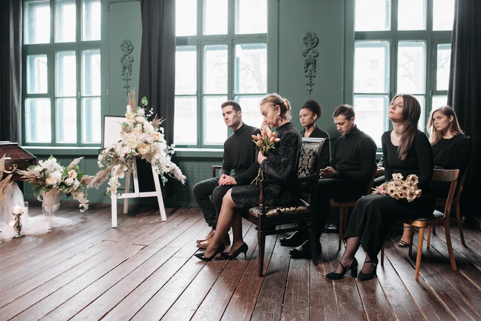 A group of six people dressed in dark clothing seated indoors near large windows with natural light, attending a somber event. The woman in the center holds a bouquet of tulips and roses, with her eye