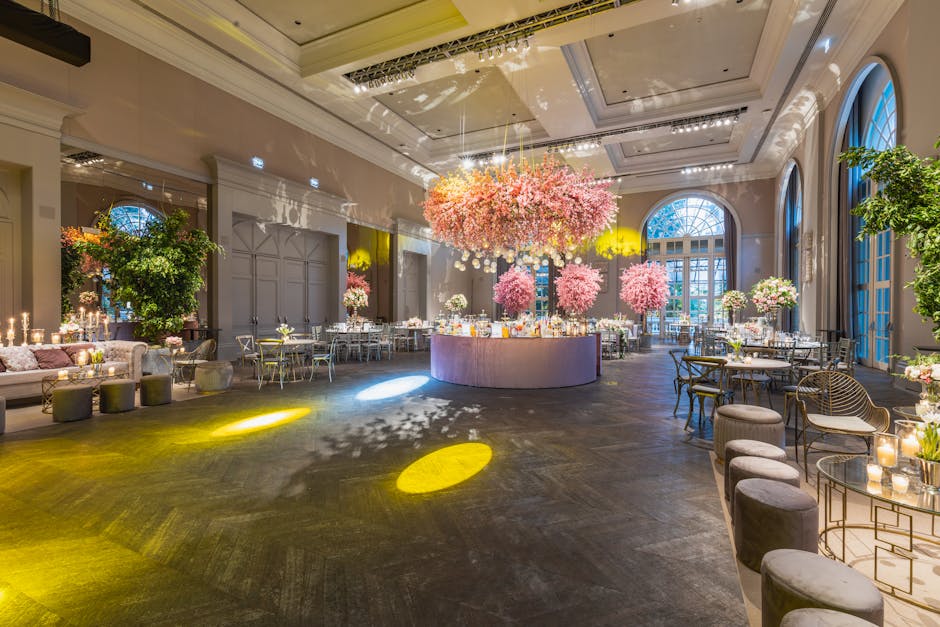 An elegant interior space featuring a grand staircase adorned with cascading floral arrangements of pink and white roses, peonies, and greenery, which wind along the banister. In the background, a lar