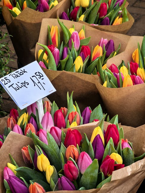 A vibrant display of fresh cut flowers including pink tulips, lilacs, and purple hyacinths arranged in neat bouquets. The flowers are wrapped in clear cellophane and presented on a floral stand, indic